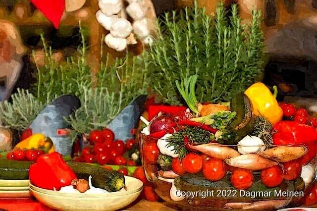 Auf einem Tisch stehen Schalen und Teller mit buntem Gemüse wie roten Tomaten und Paprika, grünen Zucchini, weißem Knoblauch und Zwiebeln. Den Hintergrund bilden Töpfe mit frischem Rosmarin.